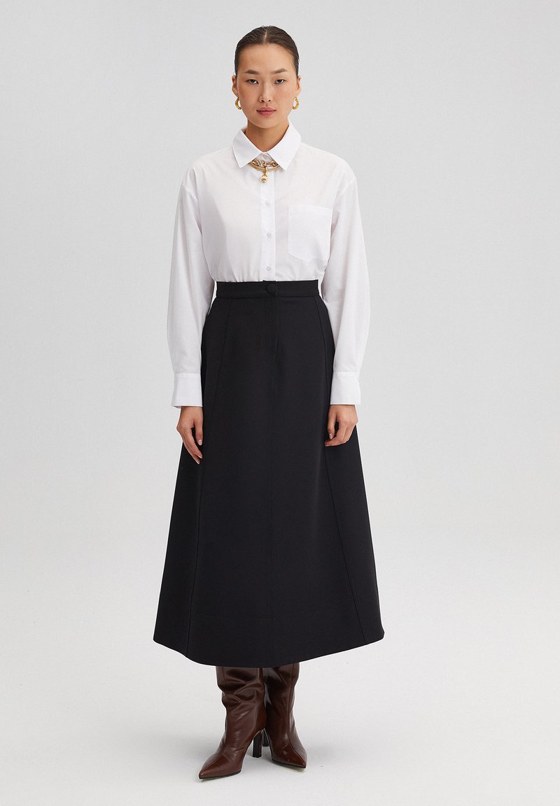 White collared shirt with a pocket, paired with a high-waisted black maxi skirt. Brown knee-high heeled boots complete the outfit.
