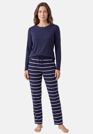 Woman standing barefoot wearing navy long-sleeve top and navy pants with white horizontal stripes and a drawstring waist.