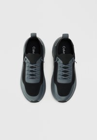 Sorte og grå treningssko med mesh-overdel, grå snørebånd, polstret krage og gummisåle. Har logo på innersålen.