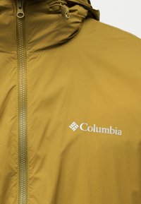 Μουσταρδί κίτρινο μπουφάν Columbia με εμπρόσθιο φερμουάρ και ρυθμιζόμενο κορδόνι κουκούλας, εστιασμένο στο λογότυπο της μάρκας στο στήθος.