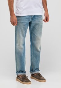 Hellblaue Jeans aus Denim mit geradem Bein, versehen mit ausgewaschenen Stellen und umgeschlagenen Säumen. Kombiniert mit braunen Wildleder-Bootsschuhen.