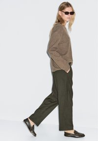 Bruine gebreide trui, olijfgroene wijde broek en bruine instappers; model loopt, draagt donkere zonnebril tegen een lichte achtergrond.