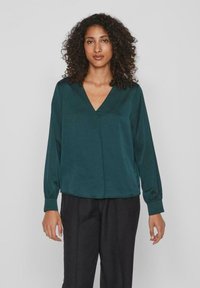 Blusa di seta verde acqua con scollatura a V e maniche lunghe, caratterizzata da una vestibilità rilassata e una superficie liscia. Abbinata a pantaloni neri.