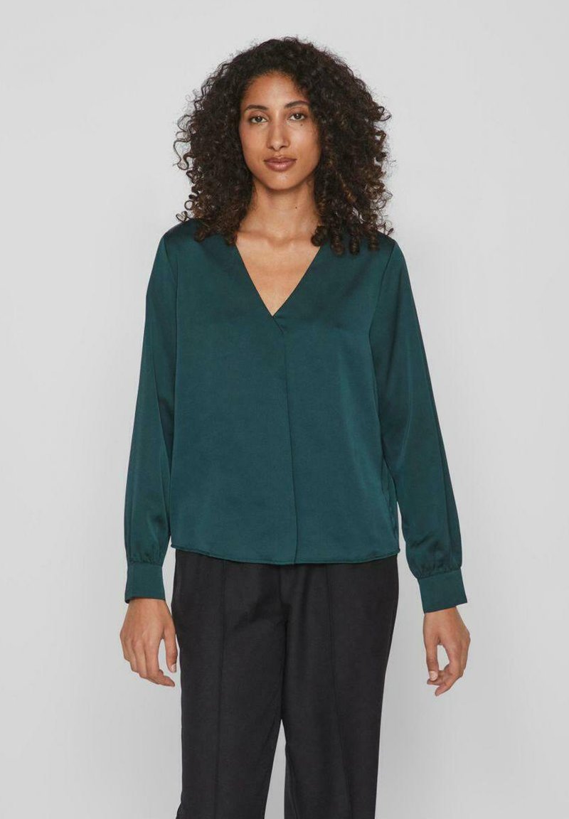 Blusa di seta verde acqua con scollatura a V e maniche lunghe, caratterizzata da una vestibilità rilassata e una superficie liscia. Abbinata a pantaloni neri.