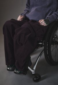 Pantaloni din catifea in violet inchis, asortați cu teniși texturați de culoare închisă. Mâinile vizibile, așezate pe un braț de sprijin al scaunului cu rotile.