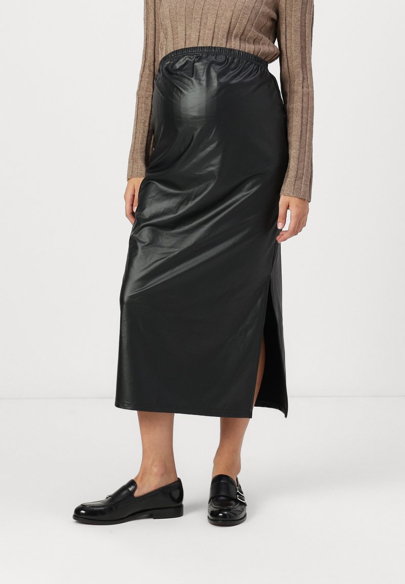 Jupe midi en simili cuir noir avec une taille élastique, dotée d'une fente sur le côté et d'une texture lisse et brillante. Associée à des mocassins noirs.
