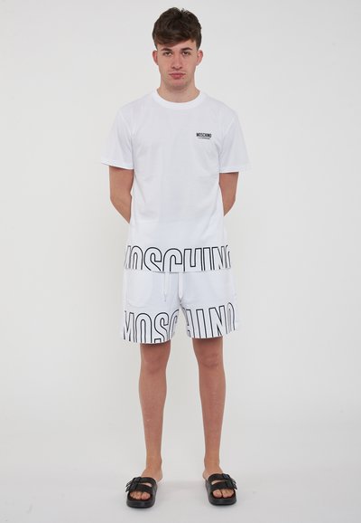 Maglietta di cotone bianca con logo nero sul petto, abbinata a pantaloni corti bianchi con scritte nere e cordoncino, indossata con sandali neri.