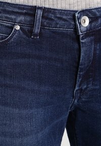 Mörka denimjeans med en slät textur, femficksdesign, metallknäppning och ton-i-ton söm detaljer längs sömmarna.