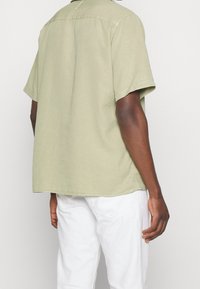 Chemise à manches courtes en tissu vert olive clair, dotée d'un col standard et d'une coupe décontractée avec un ourlet légèrement arrondi. Design simple.