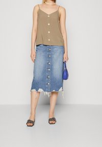 Žena v béžovém topu na knoflíky, distressed midi sukni z denimu s knoflíky vpředu, černých sandálech, s náhrdelníkem ze schránek a držící malou modrou kabelku.