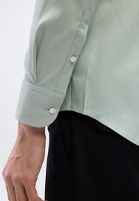 Μανίκι ανοιχτόγριου πουκάμισου με κουμπιά και μανσέτα και μπράτσο, που καλύπτει μερικώς μαύρο παντελόνι  σε έναν άτομο που στέκεται.