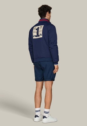 Man facing away wearing navy jacket with large "1" and Hackett London patch on back, navy shorts, white socks, and white sneakers.