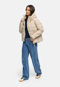 Beige vadderad jacka med huva, framsidan med dragkedja och snördetaljer, kombinerad med blå jeans och svarta och beige sneakers.