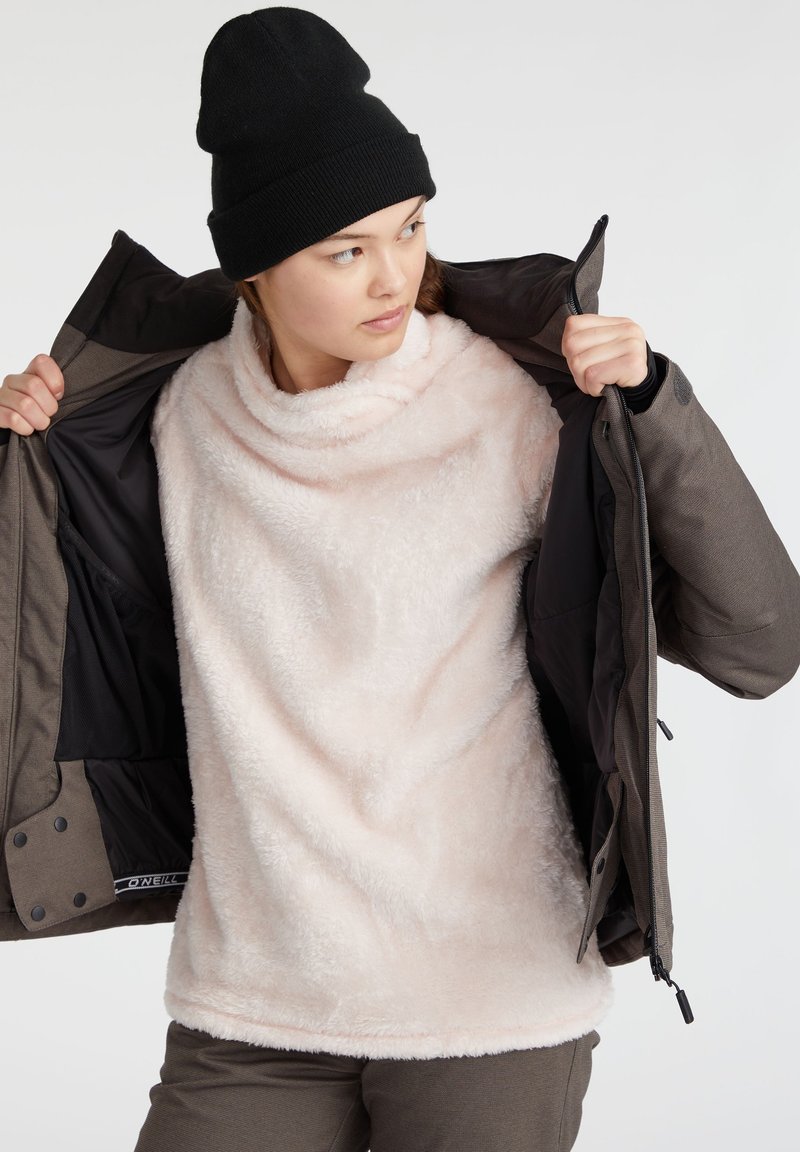 Helles rosa Fleece-Pullover mit einem Kragen in Schlauchform, getragen unter einer dunklen braunen Reißverschlussjacke. Der Pullover besteht aus weichem, strukturiertem Stoff.