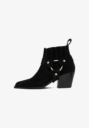 Schwarzer Wildleder-Stiefelette mit mittelhohem Blockabsatz, elastischen Seitenteilen, Riemen mit silbernem Ring und Nietenverzierung sowie Schlaufe an der Rückseite.