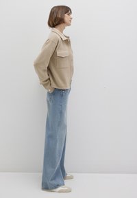 Beige wool jacket with a button-up front and two patch pockets, paired with light blue wide-leg jeans. Model stands sideways against a neutral background.