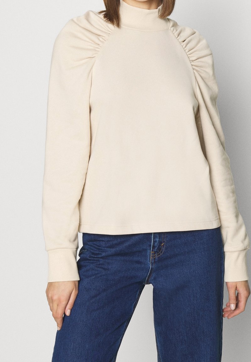 Maglione beige con collo alto e maniche a sbuffo. Tessuto morbido con una consistenza liscia. Abbinato a jeans in denim blu scuro.