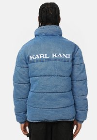 Puffer jacket in blue denim with a ribbed collar. Features horizontal stitching and "KARL KANI" embroidered in white on the back.