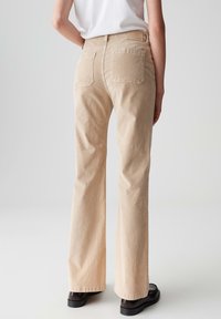 Persona che indossa pantaloni beige a gamba dritta e scarpe marroni scure, in piedi davanti a uno sfondo grigio chiaro uniforme.