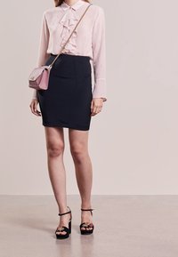 Hellrosa Bluse mit gerafftem Frontteil und langen Ärmeln, kombiniert mit einem taillierten marineblauen Rock. Schwarze Plateau-Sandalen mit floralen Verzierungen.