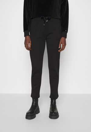 Personne portant un pantalon noir ajusté avec une taille à cordon et des bottes noires à semelle plateforme épaisses, debout devant un fond clair uni.