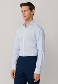 Muž v světle modré košili s knoflíky a tmavě modrých kalhotách stojící před jednobarevným světlým pozadím a dívající se stranou.