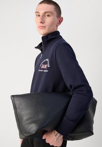 Black leather bag with a smooth texture, angled design, held under the arm. Model in a navy pullover with a zipper and white graphic print.