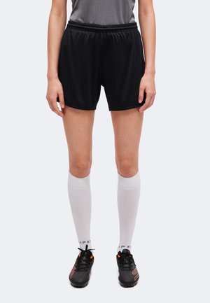 Personne portant un short de sport noir, des chaussettes blanches hautes jusqu'aux genoux, des crampons noirs et un haut gris, debout devant un fond uni.