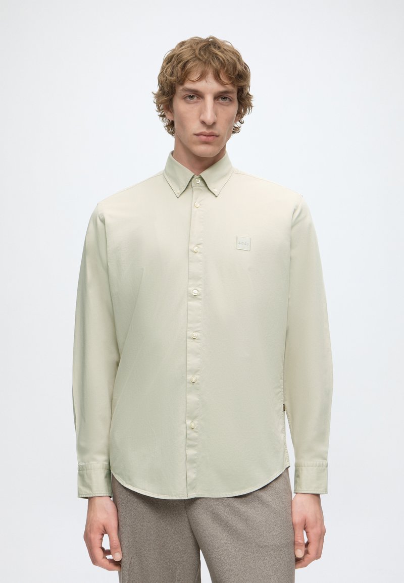 Jeune homme aux cheveux bouclés portant une chemise beige clair à boutons et un pantalon gris, debout devant un fond blanc uni.