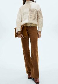 Cremfarbener Strickpullover mit strukturiertem Patchwork, kombiniert mit braunen Cordhosen im Schlagstil und braunen Schuhen. Enthält eine passende braune Tasche.