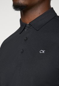 Juodas polo marškinėliai su tekstūruotu audiniu, trimis mygtukais ir baltu "CK" logotipu ant krūtinės. Yra klasikinio apykaklės dizainas.