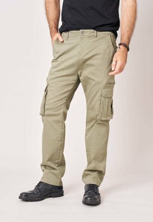 Man draagt beige cargobroek, zwarte veterlaarzen, zwart overhemd, staat met één hand in de zak tegen een effen achtergrond.