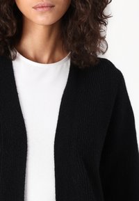 Svart ribbestrikket cardigan med åpen front, med et teksturert strikkemønster. Bærer over en hvit rundhalset topp.