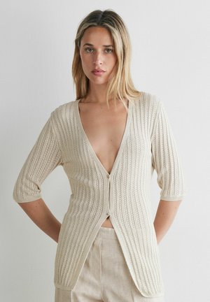 Kvinde iført en beige, ribstrikket cardigan med trekvartærmer og to knapper, kombineret med lysebeige bukser, mod en ensfarvet baggrund.