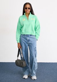 Felpa cropped verde menta con colletto a zip, abbinata a jeans cargo blu chiaro con molte tasche. Porta una borsa nera.