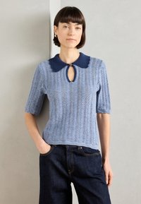 Ανοιχτό μπλε πλεκτό τοπ κοντομάνικο με ναυτική κολάρο, λεπτομέρεια κλειδαρότρυπας και υφασμάτινο σχέδιο. Συνδυασμένο με σκούρες τζην παντελόνες.