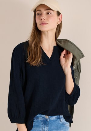 Femme portant une casquette beige, un haut à manches longues noir texturé, un jean bleu clair, tenant une veste verte sur l'épaule devant un fond beige.