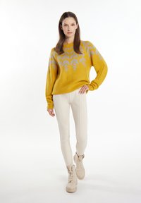Maglione giallo senape con motivo a foglie in grigio chiaro, abbinato a pantaloni aderenti color crema e stivaletti beige con lacci.