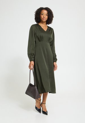 Femme portant une robe midi vert foncé à manches longues avec des talons noirs, tenant un sac à main tressé marron foncé sur un fond uni.