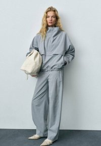 Grijze outfit met een ritssluiting, oversized jas en wijde broeken, beide in vergelijkbare stof. Model houdt een lichtgekleurde tas vast.