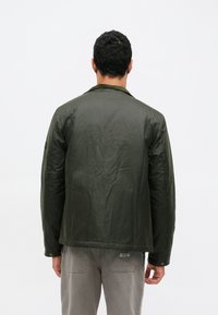 Jachetă matlasată verde măsliniu, cu guler și manșete cu nasturi, având o textură netedă și o croială lejeră. Branding subtil pe mânecă.