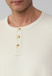 Beżowa, prążkowana koszulka henley z okrągłym dekoltem i czterema kontrastującymi żółtymi guzikami na plisce. Miękka bawełniana struktura.