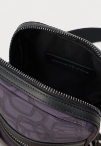 Violet spalvos per petį nešiojama rankinė su juodu odiniu dekoru, atidarytas pagrindinis skyrius rodo juodą audinį viduje ir užtrauktuką.