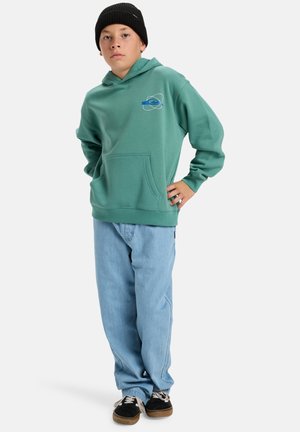 Grüne Fleece-Hoodie mit Fronttasche, Logo-Design in Blau und Weiß; kombiniert mit lockeren hellen Jeans und schwarzen Schuhen.