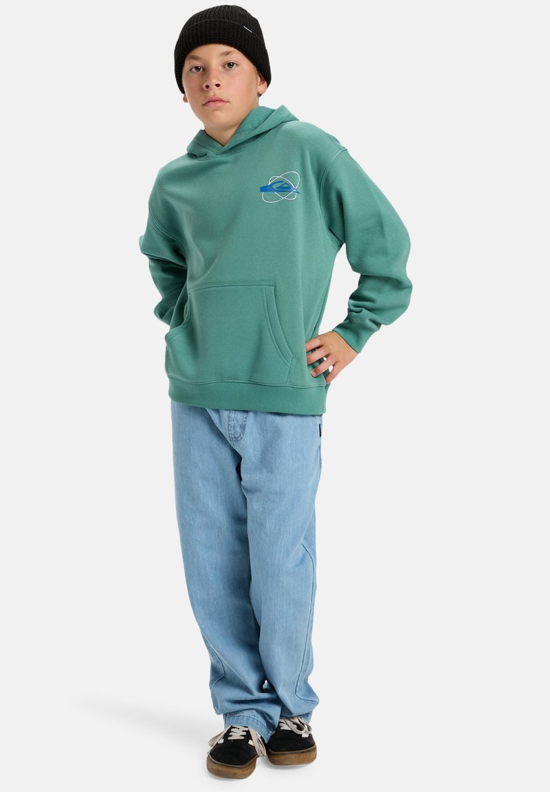 Grön fleecehuva med framficka, logotypdesign i blått och vitt; kombinerad med lösa ljusa jeans och svarta skor.