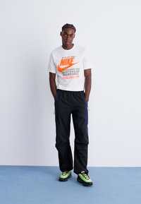 White Nike t-shirt with orange logo text, paired with black pants featuring side accents, and black and neon green sports shoes.