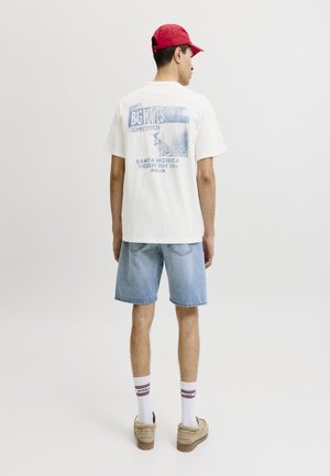 Bărbat purtând o șapcă roșie, tricou alb cu imprimeu, pantaloni scurți din denim deschis, șosete albe și pantofi bej stă cu spatele într-un studio simplu.