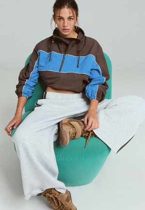 Femme avec les cheveux attachés, portant une veste marron et bleue, un pantalon large gris clair et des bottes marron, assise sur une chaise verte arrondie.