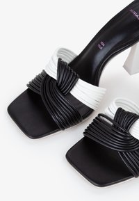 Sandali block-heel in bianco e nero con punta quadrata, caratterizzati da cinturini intrecciati in materiale sintetico e una superficie liscia.