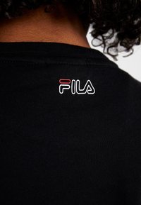 Černé tričko s malým vyšitým logem FILA v červené a bílé barvě na zadní straně. Látka vypadá měkce a má hladkou texturu.